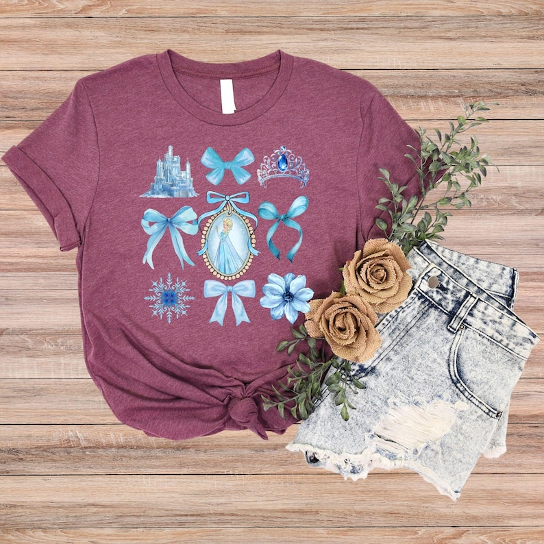 May include: Burgundy t-shirt with a watercolour design of a castle, bows, a tiara, and a princess portrait. The shirt is styled with denim shorts and flowers on a wooden surface.
