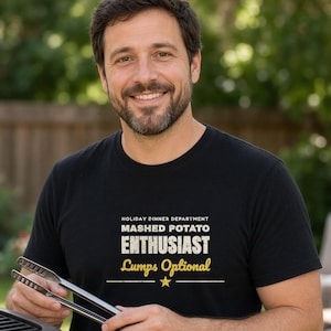 May include: A black short-sleeved t-shirt featuring white and yellow text that reads HOLIDAY DINNER DEPARTMENT MASHED POTATO ENTHUSIAST Lumps Optional with a small yellow star graphic centered below the text on the front of the garment.