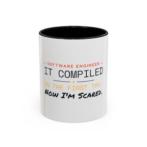 May include: A white ceramic mug with a black interior and rim. The front features the text: SOFTWARE ENGINEER IT COMPILED ON THE FIRST TRY. NOW I'M SCARED. The words are printed in a mix of orange, black, and yellow sans-serif fonts.