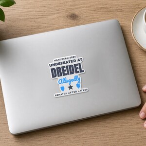 以下が含まれることがあります： シルバーのノートパソコンに貼られたダイカットステッカー。HANUKKAH 2026 UNDEFEATED AT DREIDEL Allegedly REMATCH AFTER LATKESという濃い青と水色の文字に加え、2つの小さなドレイドルのアイコンと中央に濃い青の星のアイコンがデザインされています。