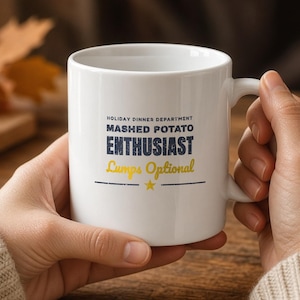 May include: A white ceramic mug featuring the text "HOLIDAY DINNER DEPARTMENT MASHED POTATO ENTHUSIAST Lumps Optional" in blue and yellow lettering, with a small yellow star and a horizontal line centered below the text.