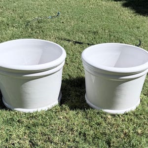 May include: Two white ceramic planters with a rounded top and a slightly flared base. The planters are sitting on green grass. The planters are suitable for indoor or outdoor use.