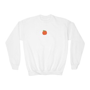 May include: A white long-sleeve crewneck sweatshirt featuring a small, centered graphic of an orange peach with a single green leaf. The garment has a ribbed collar, cuffs, and hem, providing a clean and simple look with a central fruit illustration.