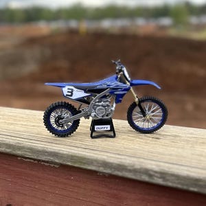 Könnte beinhalten: Blau-weißes Yamaha-Motocross-Modell mit der Nummer 3 auf der Seite. Das Modell steht auf einem schwarzen Ständer mit dem Namen "DUFFY". Das Motorrad hat schwarze Reifen und silberne Akzente.