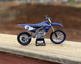 May include: A blue and white Yamaha dirt bike model with the number 3 on the side. The model is on a black stand with the name "DUFFY" on it. The bike has black tires and silver accents.