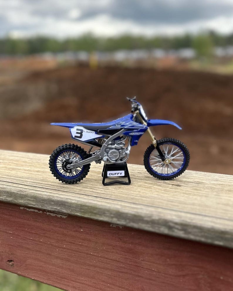 K&ouml;nnte beinhalten: Blau-wei&szlig;es Yamaha-Motocross-Modell mit der Nummer 3 auf der Seite. Das Modell steht auf einem schwarzen St&auml;nder mit dem Namen "DUFFY". Das Motorrad hat schwarze Reifen und silberne Akzente.