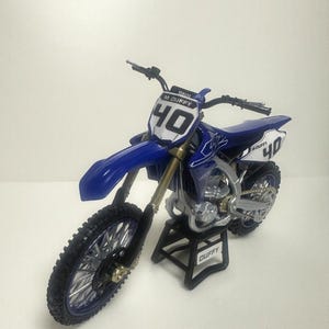 K&ouml;nnte beinhalten: Blau-wei&szlig;es Yamaha-Motocross-Motorrad mit der Nummer 40 auf der Vorder- und Seitenverkleidung. Es hat schwarze Reifen und steht auf einem schwarzen St&auml;nder mit dem Namen "DUFFY". Der Lenker ist schwarz.