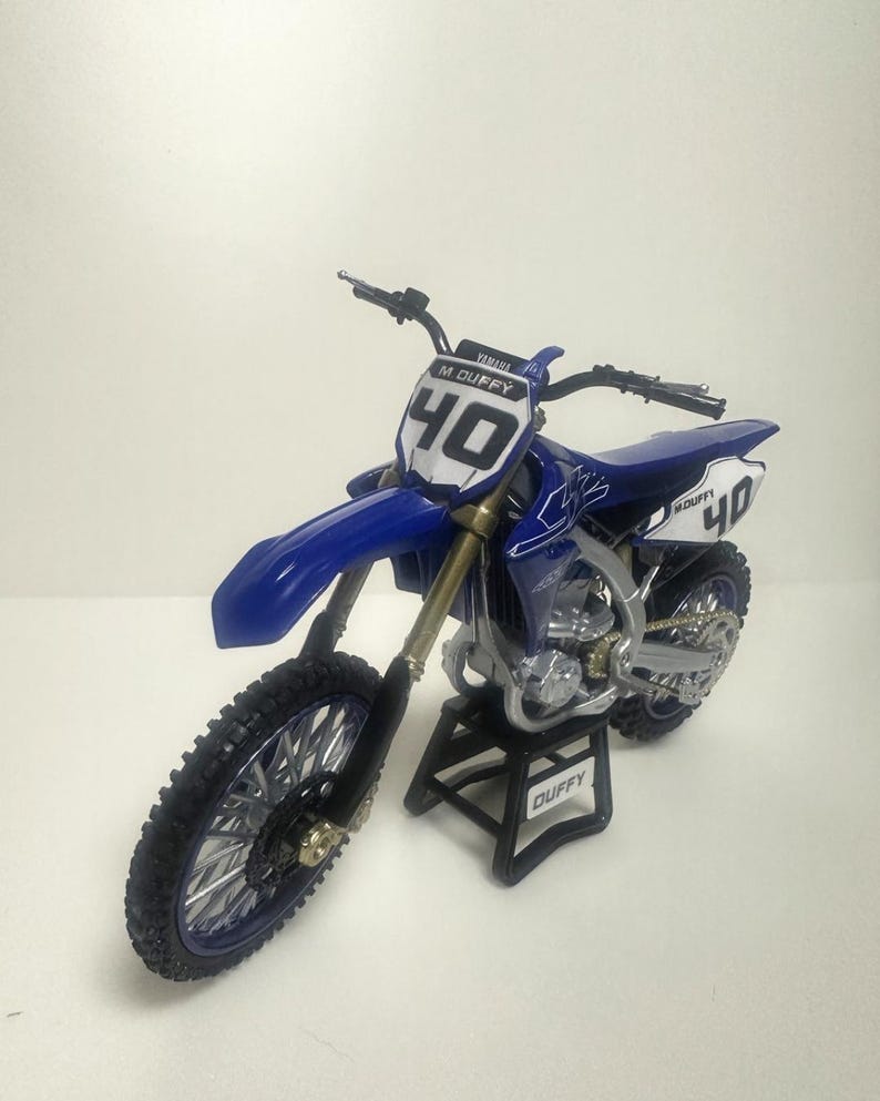 K&ouml;nnte beinhalten: Blau-wei&szlig;es Yamaha-Motocross-Motorrad mit der Nummer 40 auf der Vorder- und Seitenverkleidung. Es hat schwarze Reifen und steht auf einem schwarzen St&auml;nder mit dem Namen "DUFFY". Der Lenker ist schwarz.
