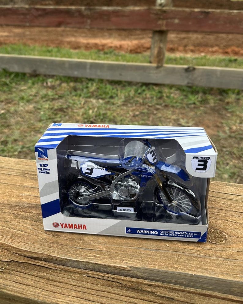 K&ouml;nnte beinhalten: Blau-schwarzes Yamaha Motorradmodell in einer transparenten Kunststoff-Vitrine. Das Modell hat die Nummer 3 und den Namen "DUFFY". Die Box tr&auml;gt das Yamaha-Logo und den Text "1:12 Die-Cast Plastic".