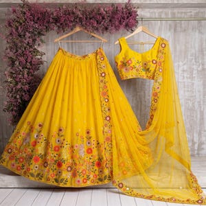 May include: A vibrant yellow lehenga set, featuring a skirt, top, and dupatta. The skirt and dupatta have floral embroidery along the edges. The top is sleeveless and also has floral embroidery. The set is displayed on wooden hangers.