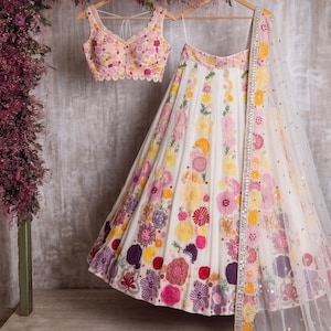 Peut inclure: Ensemble de lehenga brodé blanc avec un haut assorti et un dupatta transparent. La broderie florale présente des fleurs roses, jaunes et violettes. Le haut a un bord festonné.