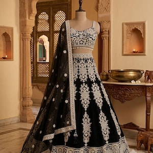 May include: A black lehenga choli set featuring intricate white embroidery. The sleeveless blouse has a square neckline with a beaded hem. The floor-length skirt has a wide embroidered border, and a matching black sheer dupatta with white floral motifs.
