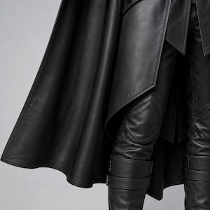 May include: A black leather garment ensemble featuring a long, flowing cape that drapes over fitted trousers. The trousers include reinforced knee panels and lower leg sections secured with horizontal buckled straps, creating a structured look.