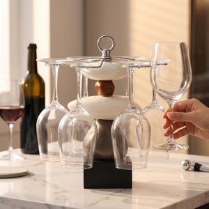May include: A wine glass rack with six clear wine glasses hanging from it. The rack is made of metal and has a decorative design. A bottle of red wine and a wine glass with red wine are in the background.