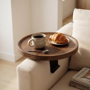 May include: A round, dark wood tray with a coffee mug, a small plate with a croissant, and a spoon. The tray is attached to the arm of a beige sofa. The mug is beige with a dark brown coffee.