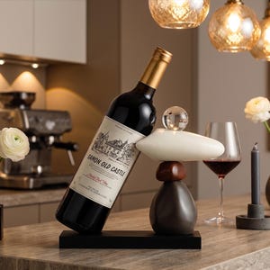 May include: A wine bottle in a modern, abstract wine holder. The holder is black with a stacked design of brown, white, and dark gray shapes, topped with a clear sphere. A wine glass and candle are also present.