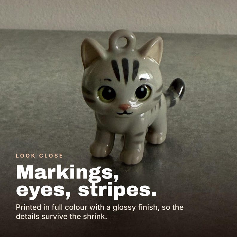 Puede incluir: Una peque&ntilde;a figura de gato de cer&aacute;mica en color gris claro brillante con rayas gris oscuro en la cabeza, el lomo y la cola. El gato tiene ojos grandes, redondos y oscuros con iris verdes, una peque&ntilde;a nariz rosa y un lazo en la parte superior.