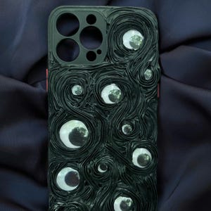 May include: Black phone case with a textured design featuring swirling lines and multiple reflective, eye-like embellishments. The case has a black camera module and a red accent on the side. The overall design is artistic and unique.