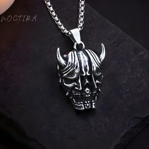 May include: A silver-colored pendant necklace featuring a detailed oni mask design. The pendant has prominent horns and intricate facial features. The necklace chain is also silver and has a box link style. The word "NOCTIKA" is in the upper left corner.