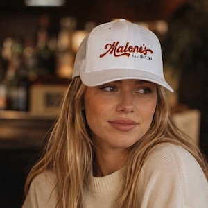 May include: A white trucker hat featuring a mesh back and a solid front panel. The front displays the red embroidered text "Malone's" in a cursive script, with the smaller black text "HASTINGS, MA" positioned directly underneath the script.