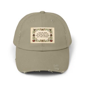 May include: A khaki distressed baseball cap featuring a rectangular patch with a botanical border of insects and flowers. The text reads: "call me when the bugs don't die and the spring looks just like autumn". The cap has a worn, frayed brim.