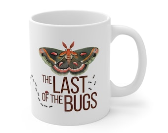 Puede incluir: Taza de cerámica blanca con una colorida ilustración de polilla con detalles en verde, rojo y marrón. El texto "THE LAST OF THE BUGS" está impreso debajo del diseño de la polilla. Una línea punteada con pequeñas ilustraciones de hormigas está a la izquierda.