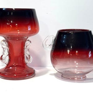 May include: Two glass vases with a red to black gradient. The taller vase has a flared top and decorative clear glass accents. The shorter vase is round with two clear handles. Both vases are on a white surface.