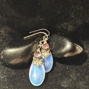 Puede incluir: Pendientes colgantes plateados con una piedra en forma de lágrima azul claro. Los pendientes tienen una parte superior plateada decorativa con una pequeña piedra rosa. Se muestran sobre una superficie oscura y texturizada.