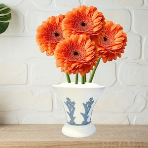 Op de afbeelding: Een boeket van levendige oranje gerbera's in een witte vaas met blauwe bloemmotieven. De bloemen, met donkere centra, zijn gerangschikt in een decoratieve vaas, wat een heldere en vrolijke weergave creëert.