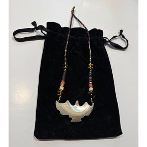May include: A handcrafted necklace with a pendant shaped like a stylized bird. The necklace features a mix of beads in brown, gold, and coral colours. It rests on a black velvet drawstring bag, creating a striking contrast.