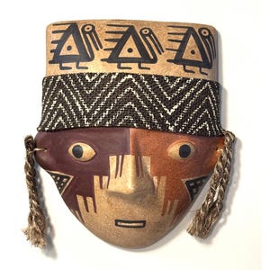 Puede incluir: Una máscara de cerámica con una cara beige, dividida en secciones marrón y naranja. Presenta diseños geométricos negros, una banda tejida y detalles de cuerda trenzada. La parte superior tiene símbolos negros de triángulo y pájaro.