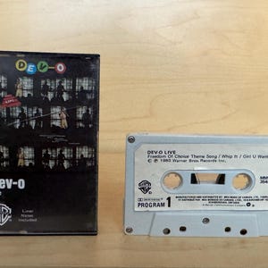 May include: A Dev-O Live cassette tape with its black plastic case. The cover features a grid of performance photos and the text Dev-O Live. The white cassette label displays the text Dev-O Live, Freedom Of Choice Theme Song, Whip It, Girl U Want, 1980 Warner Bros. Records Inc., and Program 1.