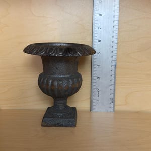 Puede incluir: Una pequeña urna gris oscura y desgastada con borde y base decorativos. La urna mide aproximadamente 13 cm de altura y se asienta sobre una base cuadrada. Una regla está colocada junto a la urna.