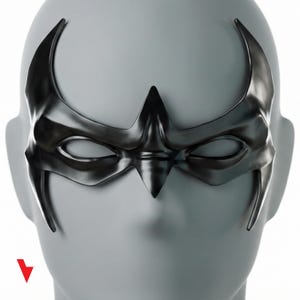 Può includere: Una maschera nera dall'aspetto metallico con un design stilizzato. La maschera presenta aperture per gli occhi e un punto centrale sopra il naso, con estensioni curve verso le tempie. La maschera è esposta su una testa di manichino grigia.