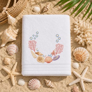 May include: A white rectangular towel featuring an embroidered design of various seashells, coral branches, and small blue bubbles arranged in a circular pattern. The towel has a textured border near the bottom edge and a soft, plush appearance.