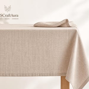 Peut inclure: Nappe en lin beige et serviette assortie sur une table en bois. La nappe présente un aspect texturé et retombe sur les bords de la table. La marque "USCraftAura" est visible en haut à gauche.