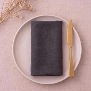 May include: A dark gray linen napkin is neatly folded on a white plate, next to a gold-colored knife. The plate rests on a pink tablecloth, with a sprig of dried flowers in the upper left corner. Table setting.