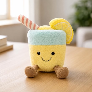 Könnte beinhalten: Ein Plüschtier in Form eines gelben Limonadengetränks mit einem hellblauen Deckel, einer Zitronenscheibe und einem gestreiften Strohhalm. Das Spielzeug hat ein lächelndes Gesicht und braune Strickfüße. Es steht auf einer Holzoberfläche.