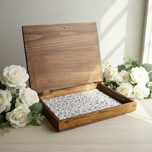 Puede incluir: Una caja de madera con tapa con bisagras, abierta para revelar un interior con estampado floral. La caja está teñida de un color marrón cálido, y la tela interior tiene un fondo blanco con pequeños diseños florales negros. Rosas blancas y vegetación están dispuestas alrededor de la caja.