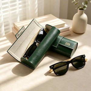 Könnte beinhalten: Ein dunkelgrünes, zylindrisches Brillenetui mit Klappverschluss und den goldgeprägten Buchstaben BB. Das Etui ist geöffnet, zeigt ein helles Innenfutter und enthält eine Sonnenbrille mit schwarzem Rahmen und hellen Bügeln aus Holz.