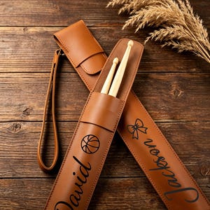 Op de afbeelding: Twee beige drumstick-etuis gemaakt van een glad materiaal, elk voorzien van een lange polsband. Eén etui is gepersonaliseerd met de naam David en een basketbalafbeelding, terwijl de andere de naam Jackson en een strik-afbeelding toont.