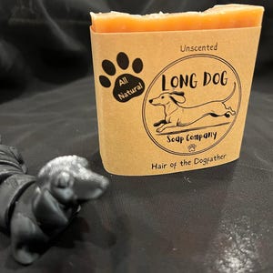 May include: A bar of unscented soap with a brown paper label featuring a dachshund illustration and the text "LONG DOG Soap Company". A small, black dachshund figurine is in the foreground.