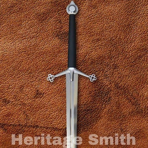 May include: A long, silver sword with a black handle and ornate crossguard. The blade is polished and reflects light. The text "Heritage Smith" is printed on the surface.