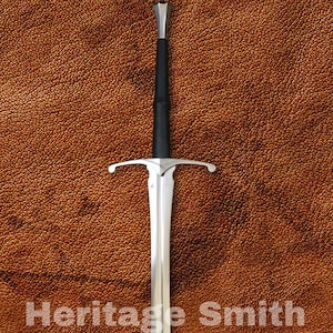 Op de afbeelding: Een lang, zilveren zwaard met een zwarte handgreep en pareerstang, tegen een bruine achtergrond met textuur. De tekst "Heritage Smith" is zichtbaar op het lemmet. Het zwaard heeft een scherp en puntig lemmet.