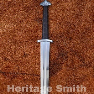 Pode incluir: Uma espada longa e prateada com punho preto e pomo decorativo. A lâmina é reta e polida, refletindo a luz. O texto "Heritage Smith" está impresso no fundo castanho.