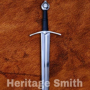 Pode incluir: Uma espada longa e prateada com punho e guarda-mão pretos, sobre um fundo castanho texturizado. O texto "Heritage Smith" é exibido em branco. A lâmina da espada é polida e reflete a luz.