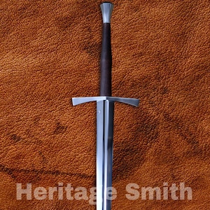 May include: A long, silver sword with a dark brown handle and crossguard, set against a textured brown background. The text "Heritage Smith" is printed on the blade. The sword has a sharp, pointed tip.