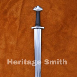 Peut inclure: Une longue épée argentée avec une poignée noire et un pommeau argenté décoratif. La lame est droite et polie, avec le texte "Heritage Smith" imprimé sur la lame. L'épée est présentée sur un fond marron.