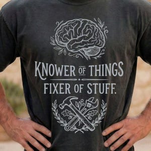 May include: A charcoal grey t-shirt featuring a white graphic print of a human brain at the top, the text "KNOWER OF THINGS FIXER OF STUFF." in the centre, and a crossed hammer and wrench surrounded by a leaf wreath at the bottom.