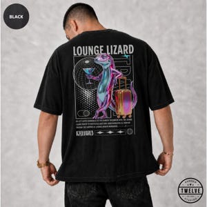 May include: A black t-shirt featuring a graphic on the back of a metallic lizard holding a martini glass next to a rolling suitcase. The design includes the text LOUNGE LIZARD, a globe grid, and small text blocks. The shirt has a loose, boxy fit.